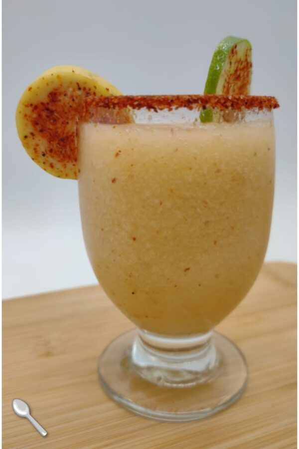 Vaso con bebida congelada frutal con borde de chile y rodajas de fruta como decoración.