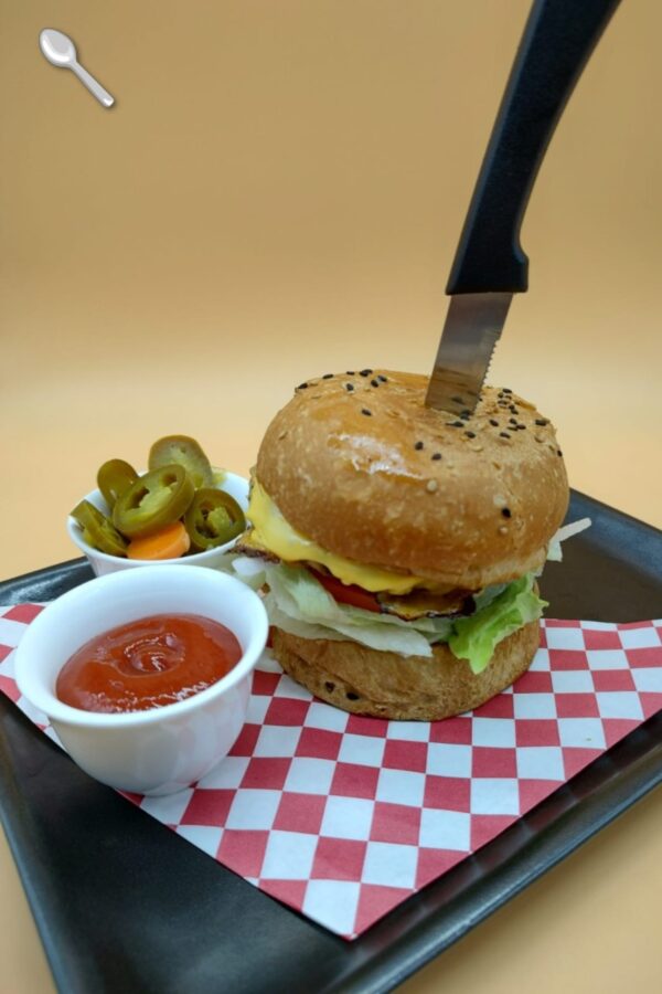 Hamburguesa artesanal con cuchillo clavado, acompañada de jalapeños y salsa en bandeja con papel cuadriculado.