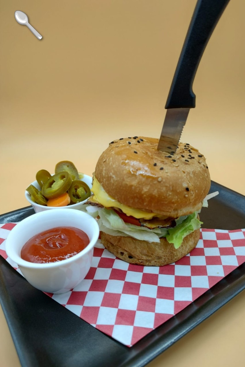 Hamburguesa artesanal con cuchillo clavado, acompañada de jalapeños y salsa en bandeja con papel cuadriculado.